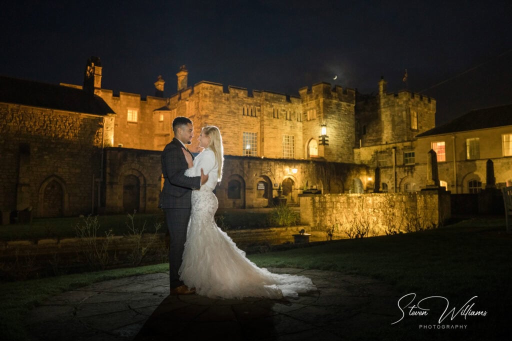 Wedding couple embracing at night before illuminated castle.