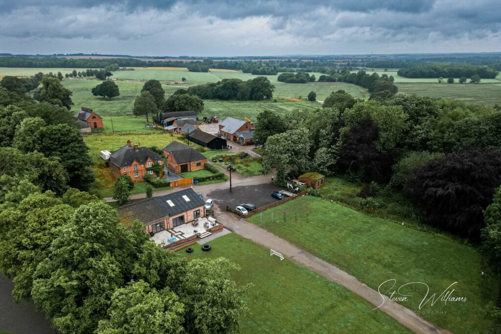 Aerial view of a rural landscape featuring a cluster of buildings with orange roofs, surrounded by lush green fields and trees, under a cloudy sky.