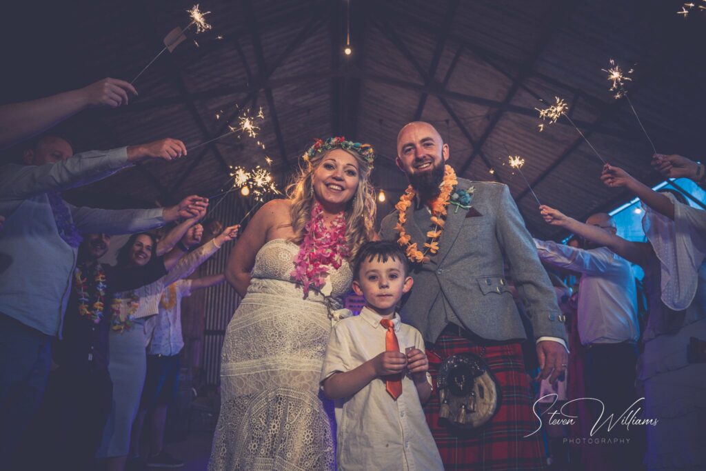 A joyful bride and groom, accompanied by a young boy, walk through a barn as guests waving sparklers surround them. the groom is in a tartan kilt and the bride wears a floral crown.