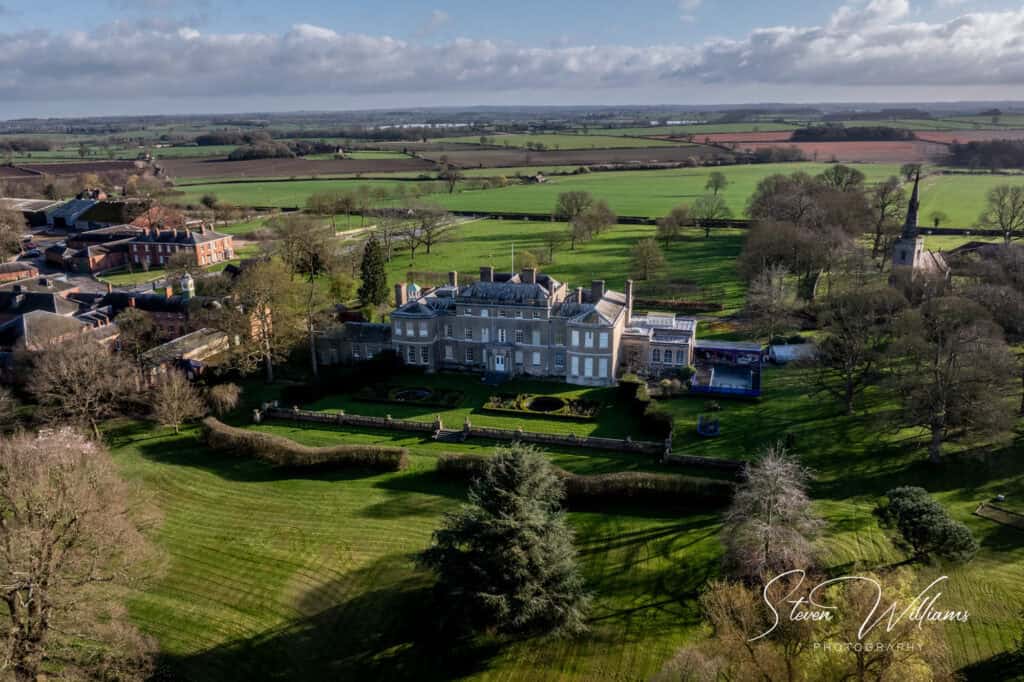 Aerial view of Thorpe Hall Estate with large, manicured gardens surrounded by lush green fields. The landscape is dotted with small clusters of buildings under a partly cloudy sky.