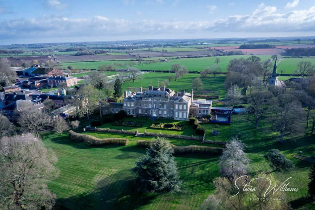 Aerial view of Thorpe Hall Estate with manicured gardens, surrounded by trees and expansive green fields, under a clear sky with scattered clouds.
