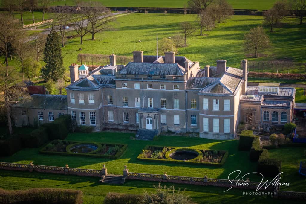 Aerial view of Thorpe Hall Estate with symmetrical gardens, featuring a grand house flanked by lush green lawns and lined pathways, under the glow of a setting sun.