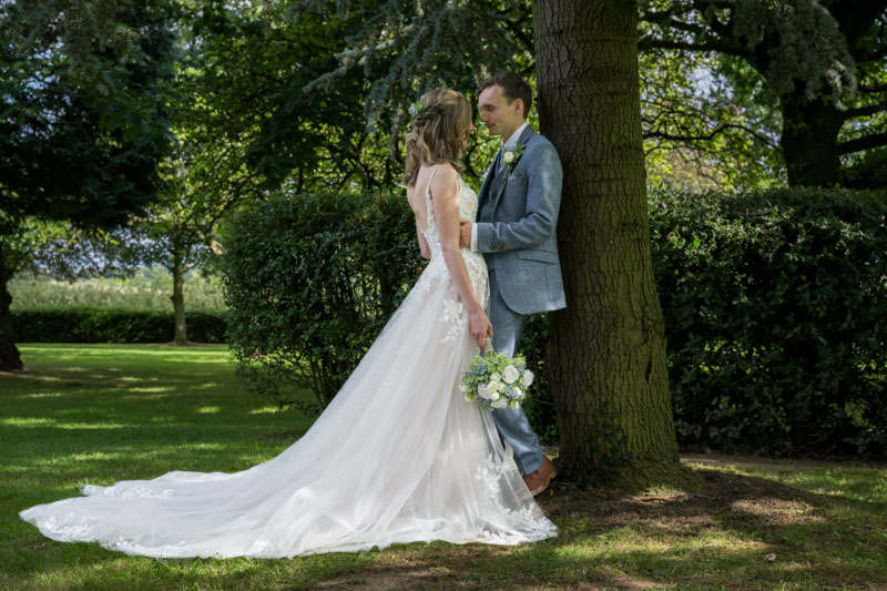 Bride and groom standing by tree in garden.