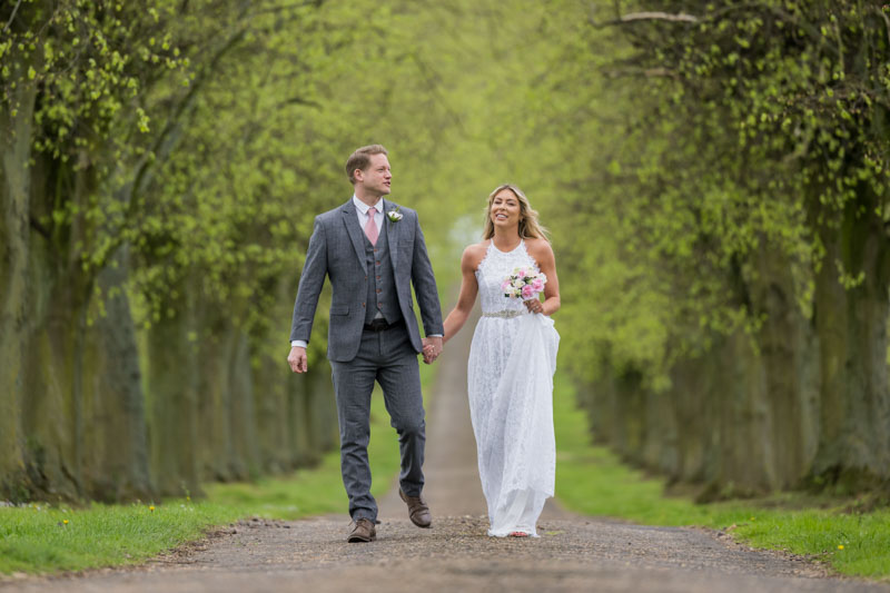 Bride and groom walking outdoors, tree-lined pathway.