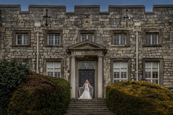 Bride on historic mansion's steps, dramatic clouds above.
