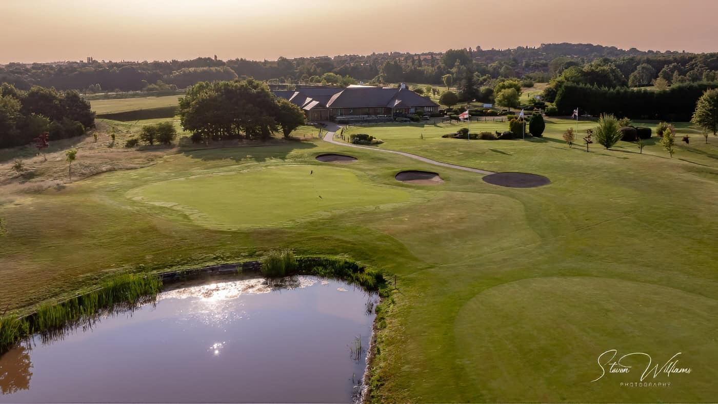 Aerial view of Aston Wood Golf Club with two bunkers and a pond in the foreground. In the distance, there is a clubhouse surrounded by trees and greenery under a golden sky. The landscape includes rolling hills and well-maintained grassy areas.
