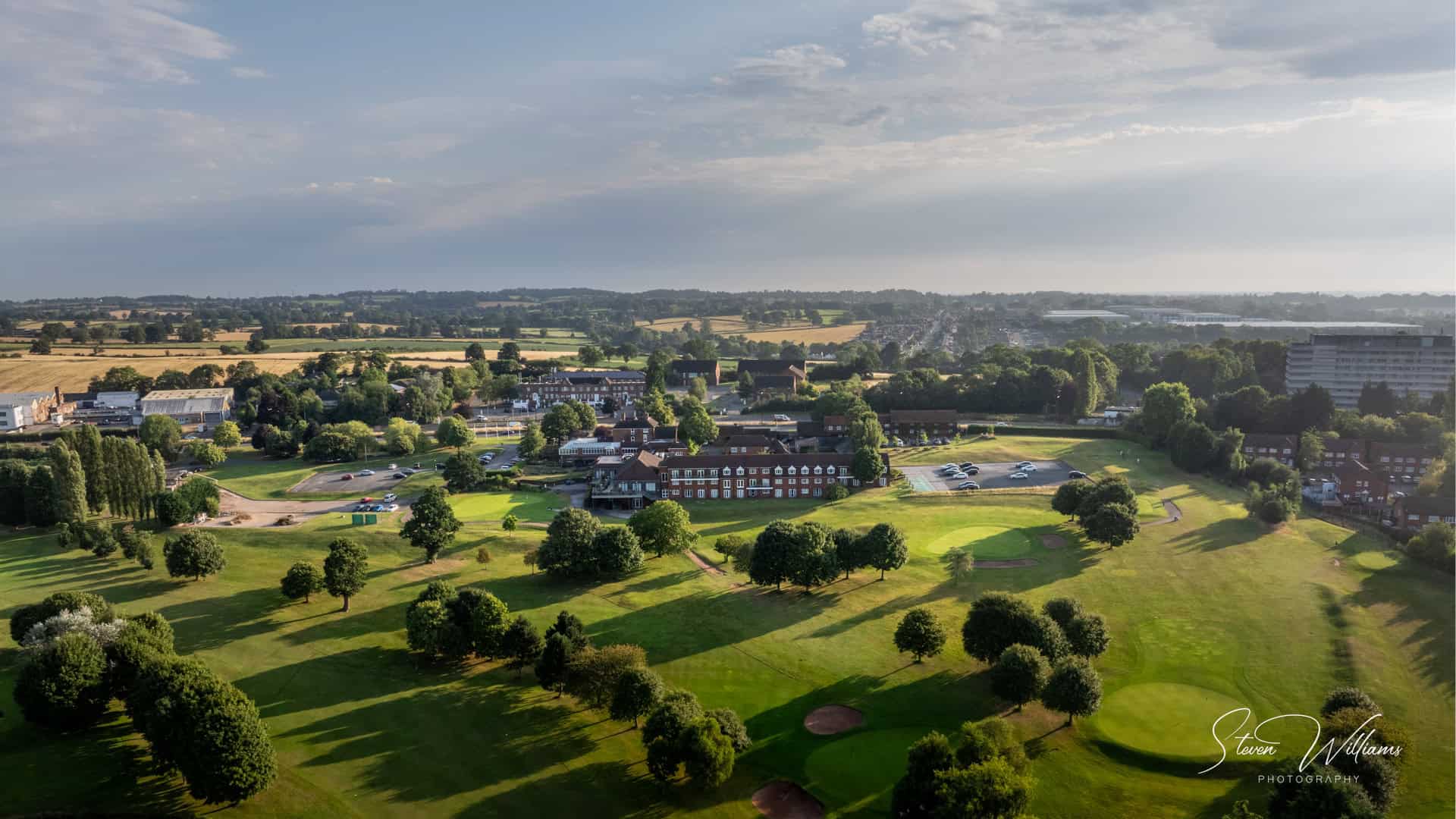 Aerial view of a lush, green golf course surrounded by trees and residential buildings. Nearby, the charming Windmill Village Hotel adds a quaint touch to the scenery. The expansive landscape extends into the horizon under a partly cloudy sky. A clubhouse and parking lot are visible in the center-right.