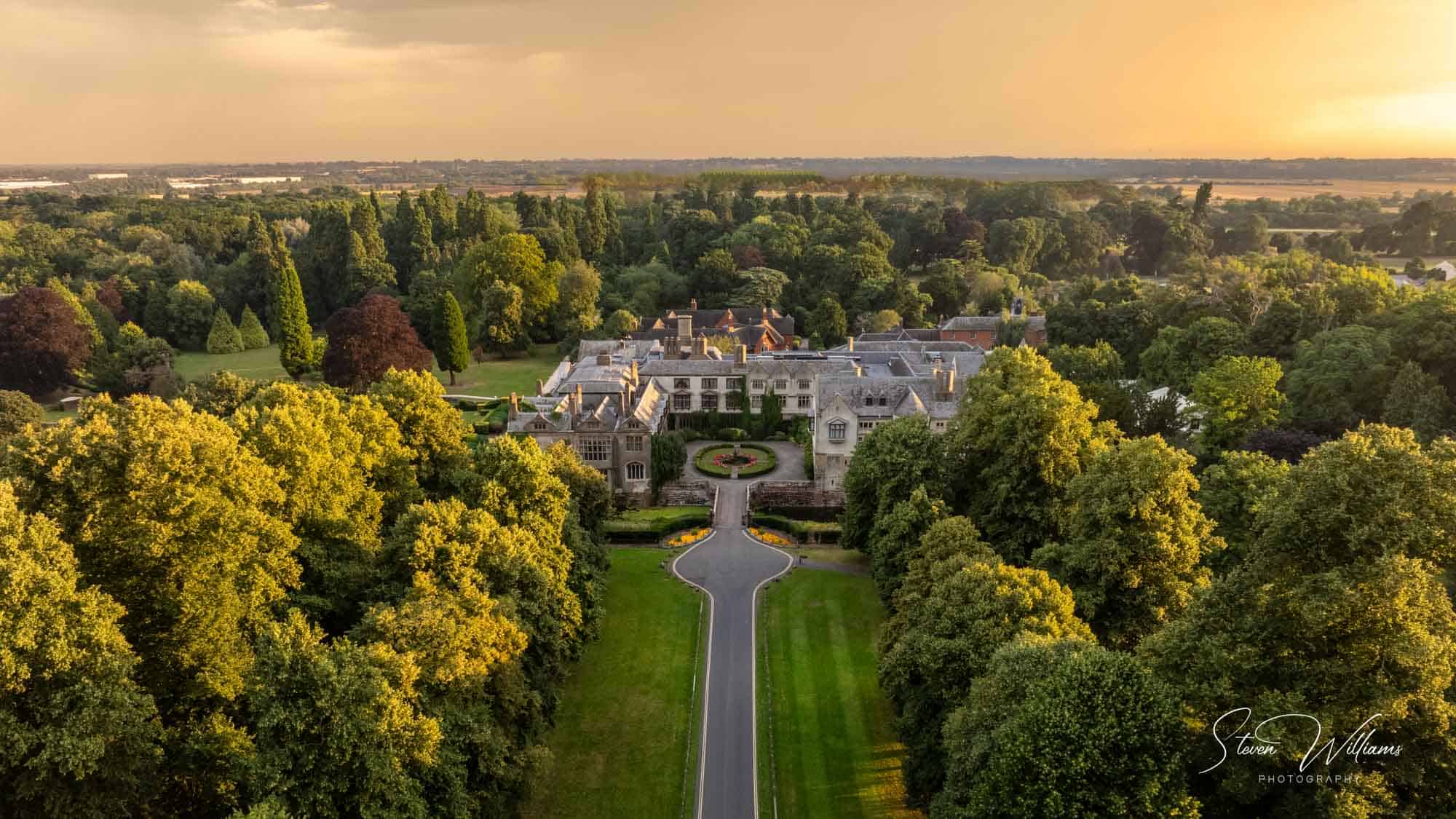 Aerial view of Coombe Abbey estate surrounded by lush trees at sunset. The central stone building is expansive with a courtyard and long driveway leading to it. The golden light creates a serene and picturesque scene. In the distance, countryside extends to the horizon.