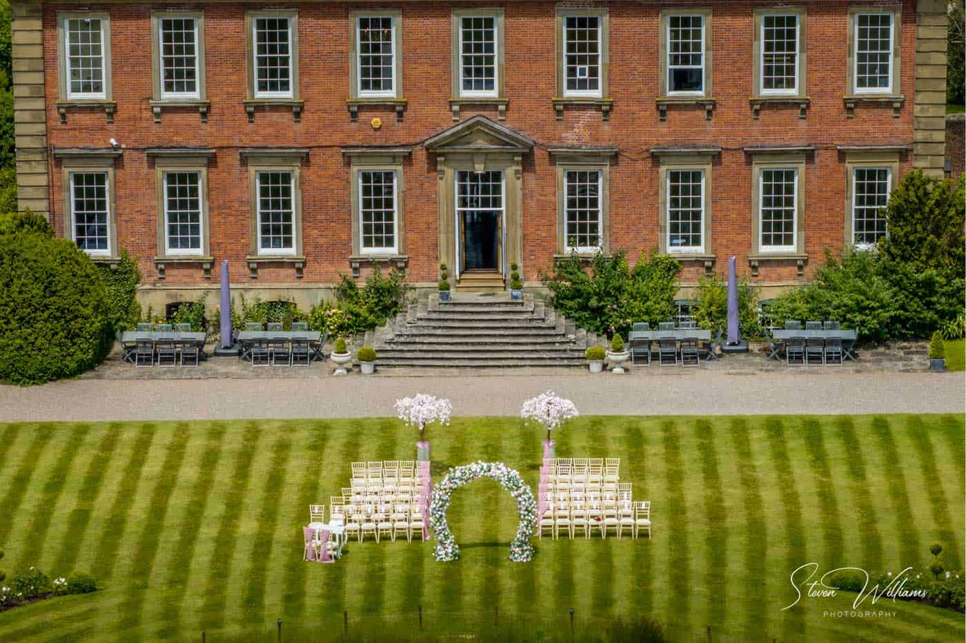 A grand brick mansion, Davenport House, serves as the backdrop for a wedding setup on a lawn. Rows of white chairs face a floral arch adorned with pink and white flowers. Symmetrical greenery and trees frame the scene, with the mansion’s windows and door centered in the background.
