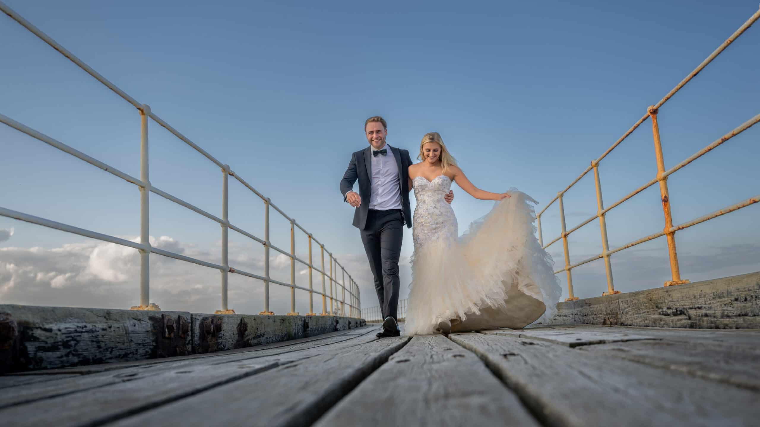 A bride in a flowing white gown and a groom in a suit joyfully walk hand in hand on Whitby pier under a clear blue sky, with the sea stretching elegantly in the background.