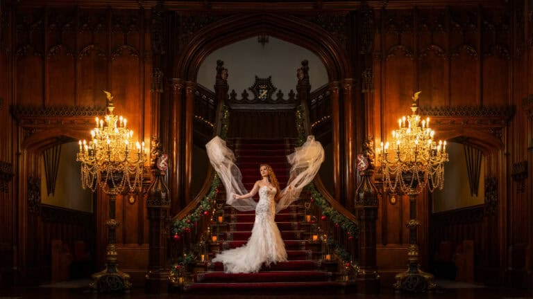 A bride in a flowing white gown stands on a grand staircase in a dimly lit, ornate hall. She raises her veil, creating an elegant curve. Two large chandeliers and intricate wooden details frame the scene, adding to the opulence.
