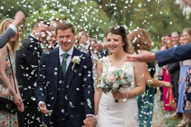A bride and groom smile and walk hand in hand through a vibrant flurry of confetti, tossed by a crowd of cheering well-wishers. The bride clutches her bouquet of flowers, both elegantly dressed in exquisite wedding attire.