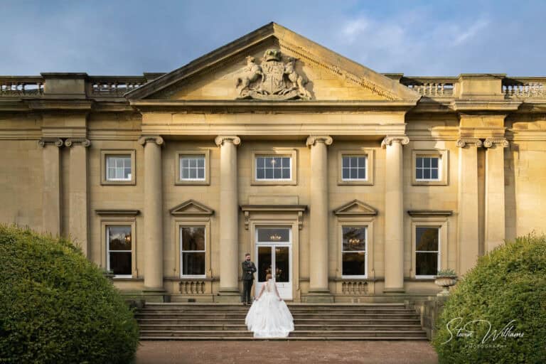 A bride in a white gown and a groom in a dark suit stand on the stone steps of Wortley Hall, a grand neoclassical building with columns and ornate carvings. The partly cloudy sky adds to the romantic backdrop, framed by lush greenery on either side.